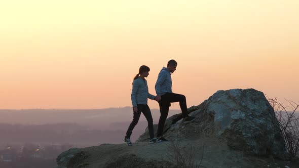 Man and woman hikers helping each other to climb stone at sunset in mountains.  alt