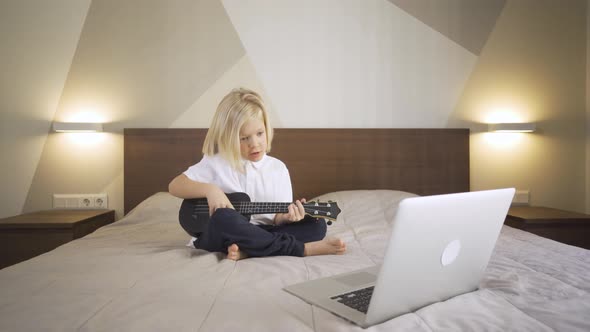 Distance Learning Online Education. A Cute Kid Learning To Play the Ukulele and Singing on a Laptop