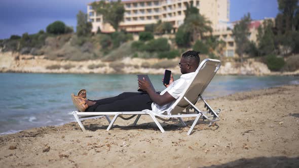 Busy African American Man Surfing Internet on Tablet Texting on Smartphone Lying on Sunbed on Sandy alt