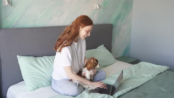 A Pregnant Woman Working at Home on a Laptop Sits on the Bed in the Bedroom and Holds Her Pet alt