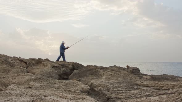 Old fisherman standing on sea side rocks and fishing against the sunset alt