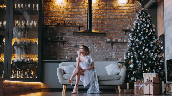 A young woman in an evening dress sits alone on a sofa on Christmas eve.	