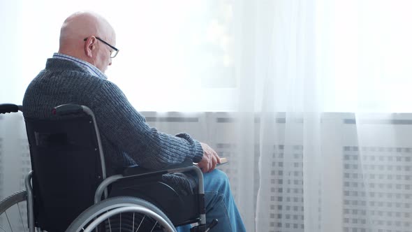 Disabled old man is sitting in a wheelchair at home alone. A handicapped person. alt