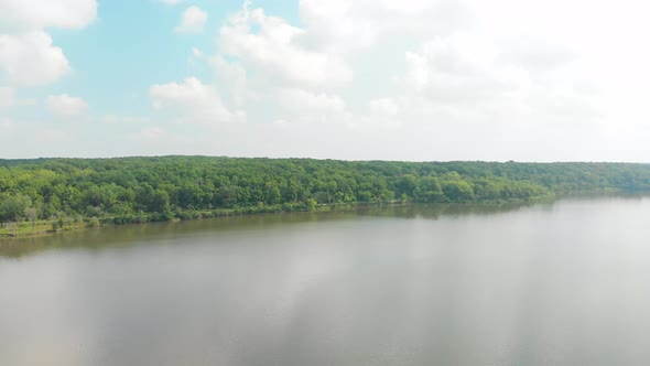 drone forward flight to a horizon and a tree line over a lake with bright green leaves with white pu alt