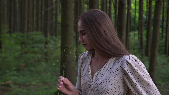 Woman in White Dress with Polka Dots Sniffing a Plucked Plant in the Forest alt