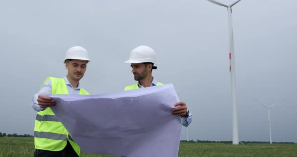 Men in white helmets hold project sheet, wind turbine generating electricity operation.