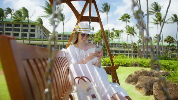 Cute Attractive Woman Swinging While Using Phone Tropical Island Resort Garden alt