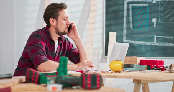 Smiling Young Craftsman Sitting at a Workbench in His Carpentry Workshop Working Online with a alt
