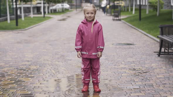 Portrait of Cute Little Girl in Pink Waterproof Costume and Rubber Boots Standing in Puddle and alt