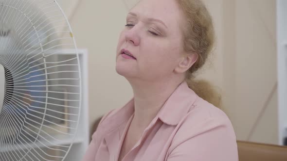 Close-up of Senior Woman Using Electric Fan Indoors. Portrait of Ill Caucasian Lady Having Fever or alt
