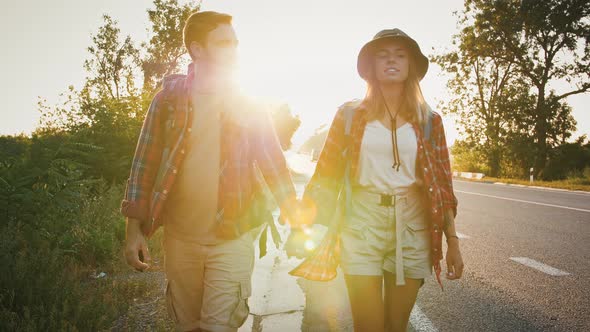 Young Couple in Love with Backpacks Holding Hands and Walking Along Road at Sunset alt