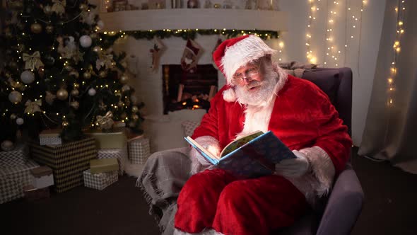 Funny Santa Clause Sitting Near Fireplace and Christmas Tree Opening a Magical Book with Shining