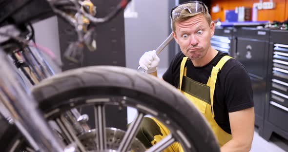 Emotionally Pensive Auto Mechanic Holds Wrench and Stares in Surprise at Motorcycle in Service alt