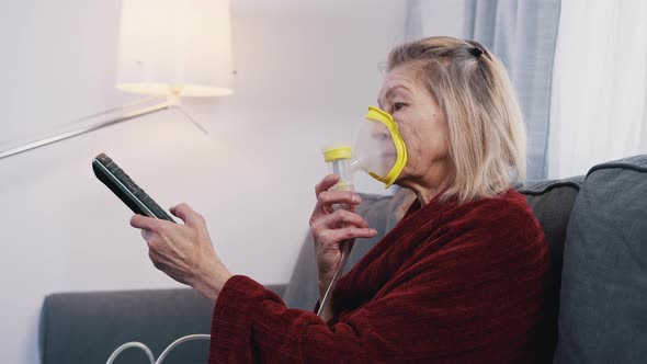 Elderly Woman Using Oxygen Mask While Changing Channels on Tv at Home alt