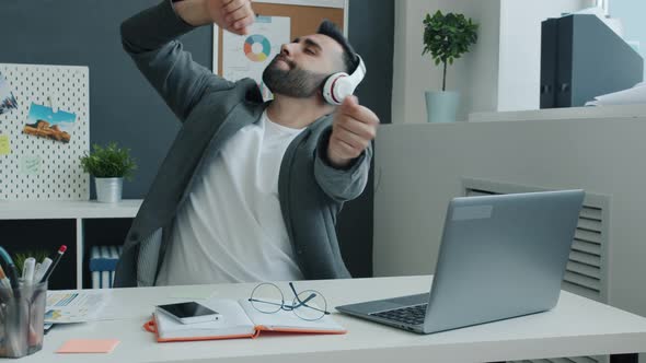 Funny Businessman Wearing Headphones Dancing and Working with Laptop Sitting at Desk in Office alt