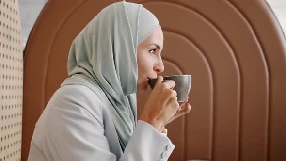 Side View of Beautiful Young Woman Wearing Hijab Drinking Coffee and Smiling in Cozy Cafe alt