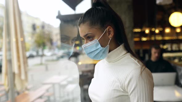 Young Woman in Face Mask Standing in Front of Windows in Cafe alt