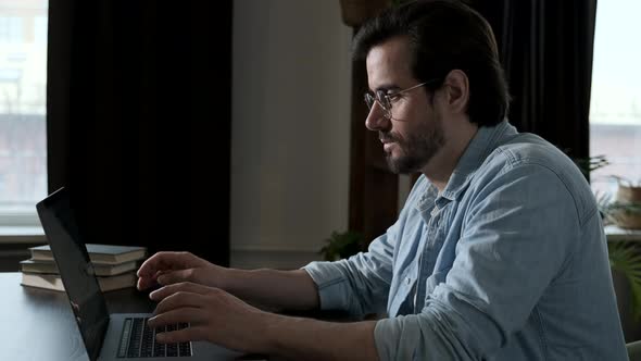 Side View of A man with Glasses, Blue Shirt and White t-Shirt Working on a Computer laptop  alt