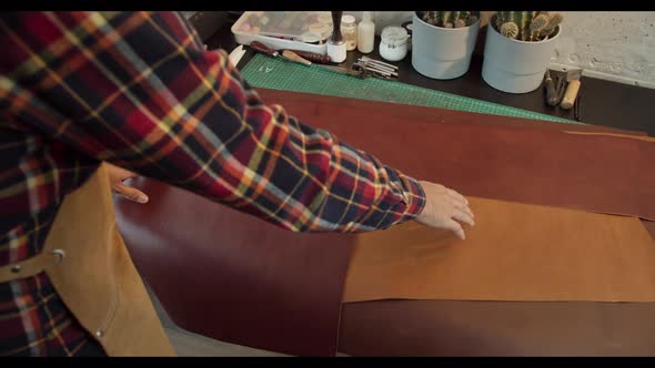 Bearded Millennial Craftsman with Mug of Hot Drink Working and Inspecting Piece of Leather While