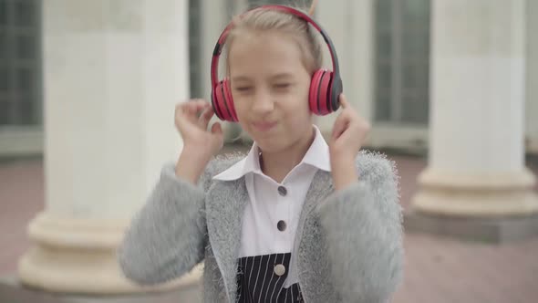 Portrait of Positive Caucasian Schoolgirl Listening To Music in Headphones Standing Outdoors. Happy alt
