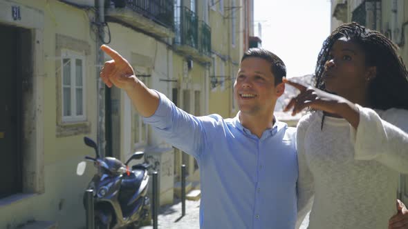 Happy Couple Hugging and Pointing Up While Walking on Street alt
