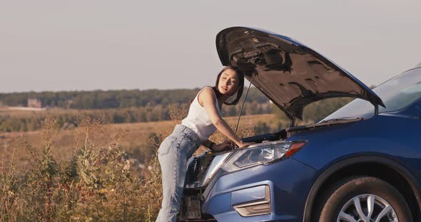 Young Woman Looking at Open Hood of Broken Car, Then Trying To Catch Vehicle for Help alt