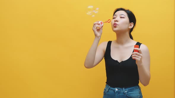 Childish Young Asian Woman Blowing Soap Bubbles. Isolated on Yellow Background alt