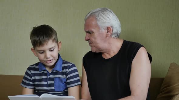 Grandson Student Reads a Book to His Grayhaired Grandfather Sitting Next to Him