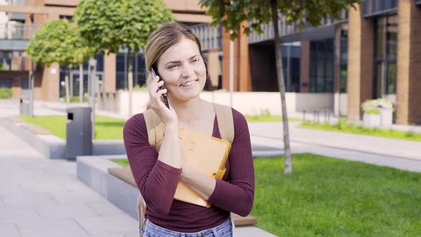 Female Student with Notebook Talks on Phone Walking Along Park