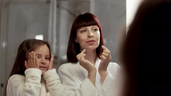 Content Young Woman and Little Daughter in White Bathrobes Applying Cream on Cheeks alt