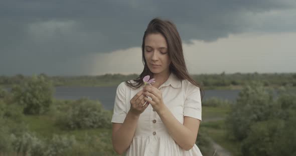 Elegant Girl in White Dress Holding Wild Flower Outdoors on Stormy Landscape Background. Long-haired alt