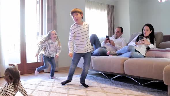 Mom and Dad Sits on Sofa and Use Phone During Their Children Play Inside alt