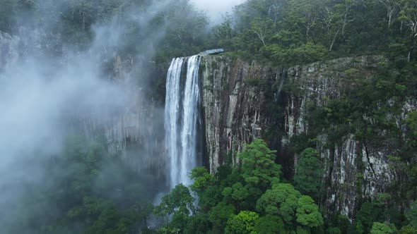 Unique drone view through suspended mist revealing a majestic waterfall spilling down a lush rainfor alt