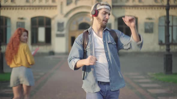 Middle Shot of Relaxed Happy Young Man in Headphones Dancing As Walking Along University Yard After alt