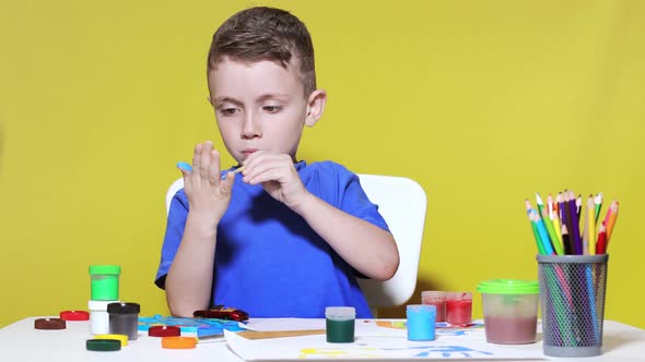 A little preschooler sits at a table, holding a brush in his hand and painting paints his palm. alt