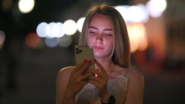 Cute Girl Uses Smartphone and Smiles Standing on Night Street in the City
