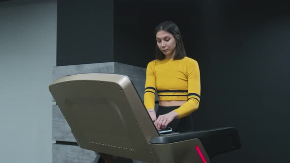 Happy Young Woman in Yellow Top Walks Warm Up on Treadmill and Puts on Headphones to Listens Music alt