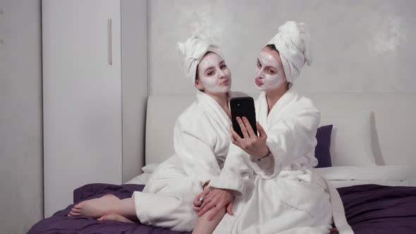 Two Girls in Bathrobes and Face Masks Take Selfies on Their Phone
