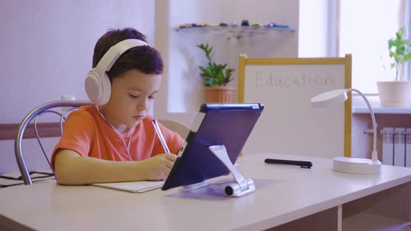 A Boy in White Headphones is Studying in an Online School on a Tablet