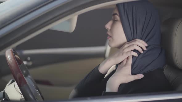 Portrait of Independent Young Muslim Woman Sitting in Her Car and Correcting Traditional Headscarf alt