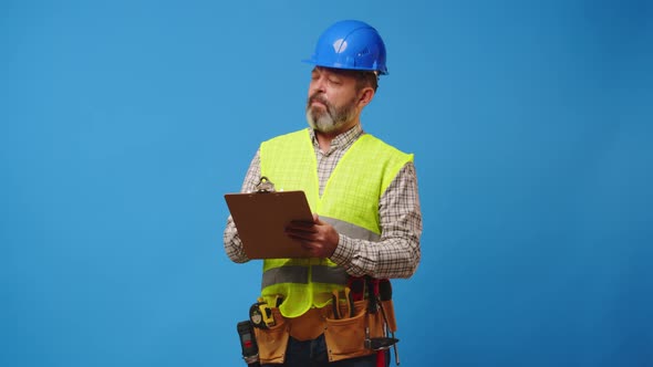 Senior Man Builder Making Notes on Clipboard Against Blue Background in Studio