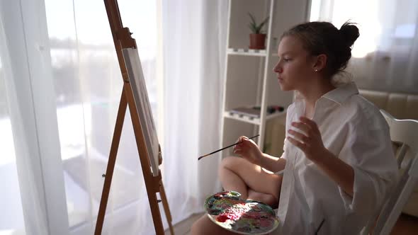 Beautiful Portrait of a Young Girl in a White Shirt Painting a Portrait on Canvas alt