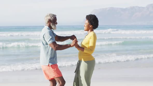 Happy african american couple dancing and holding hands on sunny beach alt