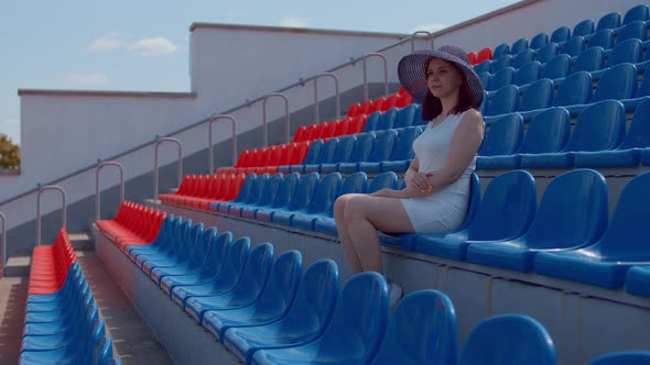 Young Woman in in White Dress and Elegant Hat Looks Away Sitting on Stadium Bleachers Alone alt
