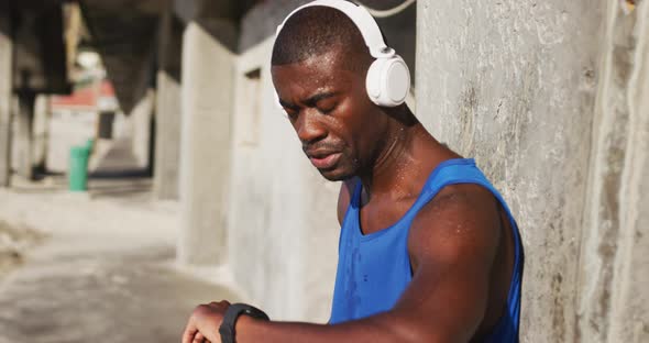 African american man wearing headphones checking his smartphone, exercising outdoors on sunny day alt
