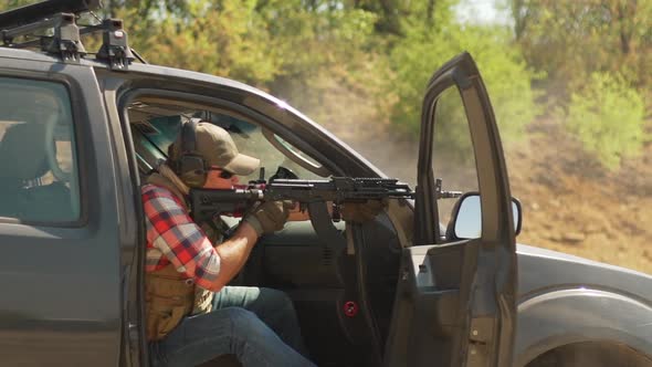 Caucasian Male in Cap Plaid Shirt and Sunglasses Firing Airsoft Gun Sitting on Car Seat alt