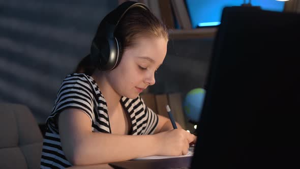 Little Girl Student Writing Homework at Night at Home Using Laptop and Headphones Elearning Online alt
