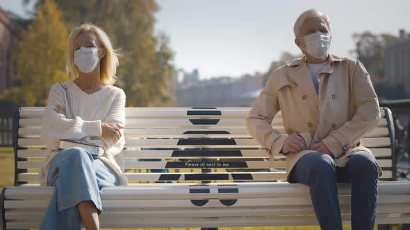 Aged Man and Woman Sitting on Bench with Keeping Social Distancing Mark Outside alt