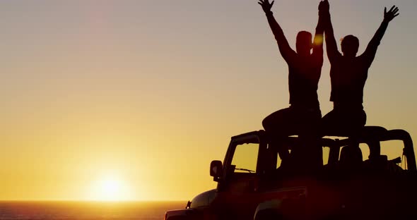 Happy caucasian gay male couple sitting on car raising arms and holding hands at sunset on the beach alt