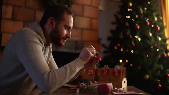 Side View of Relaxed Young Man Writing Number on Red Bag By Brush and White Paint Sitting at Table alt